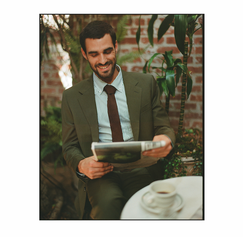 Man in an olive suit, striped shirt, and brown knit tie seated outdoors among green plants and a brick wall, smiling while reading a magazine, with a cup of coffee on a small table in the foreground.