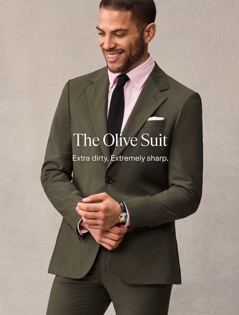 Close-up of a man in an olive suit, pink shirt, black knit tie, and white pocket square, smiling and looking down while clasping his hands, wearing a watch. Text overlaid reads: The Olive Suit. Extra dirty. Extremely sharp.