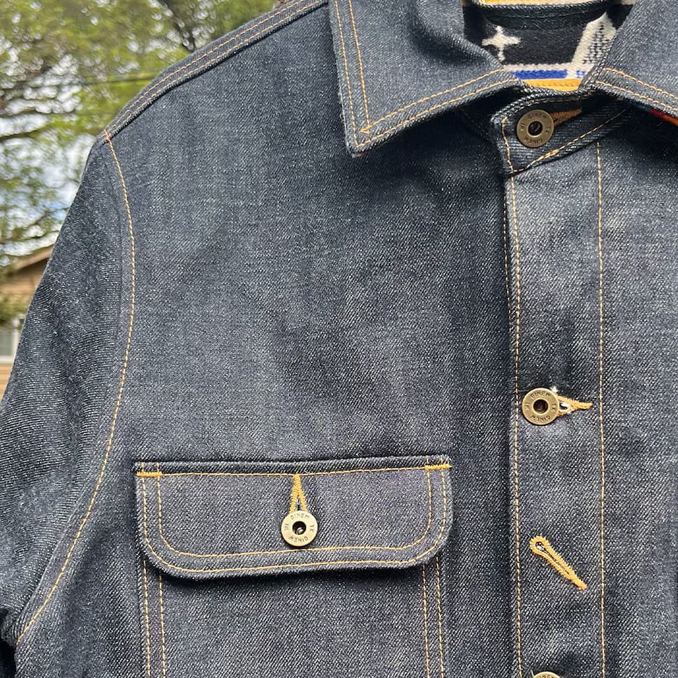 Close-up of the Bill Barn Coat's collar and chest pocket showing brass button hardware and denim stitching.