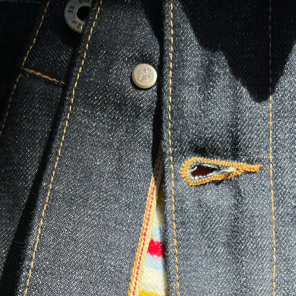 "Close-up of the Bill Barn Coat's chest pocket showing custom Ginew brass rivet hardware and topstitching detail on selvedge denim."