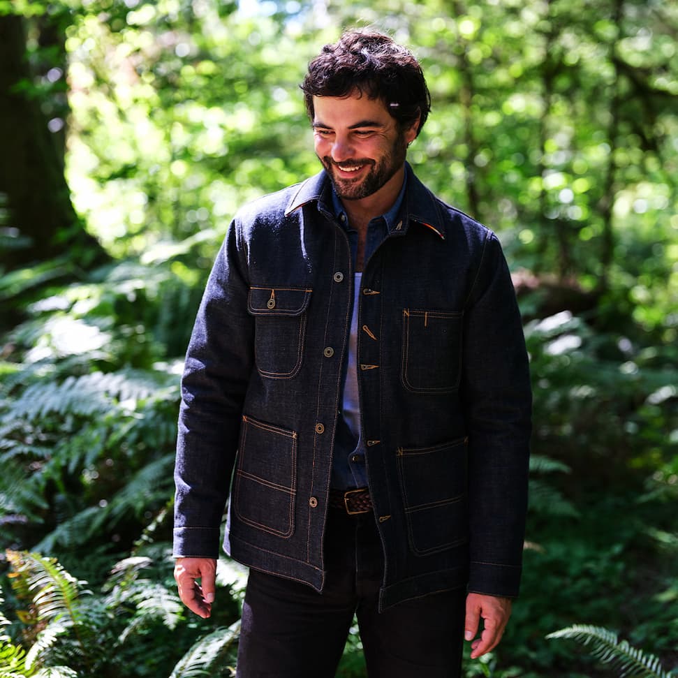 "Man wearing the Ginew Bill Barn denim coat over a collared shirt, standing in a forest"