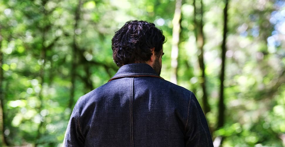 Man wearing the Ginew Bill Barn denim coat from behind, standing in a forest, showing the back yoke and stitching.
