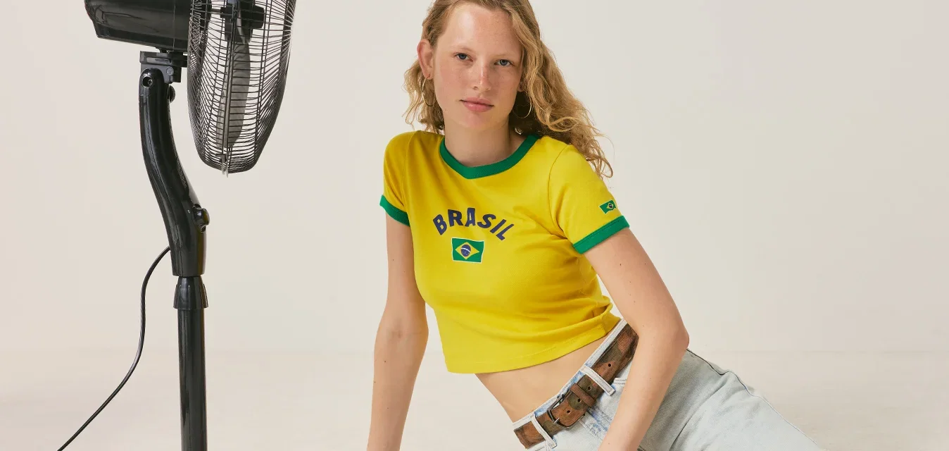 Jonge vrouw in een geel Brazilië t-shirt en spijkershorts zit op de vloer naast een zwarte staande ventilator in een eenvoudige witte kamer.