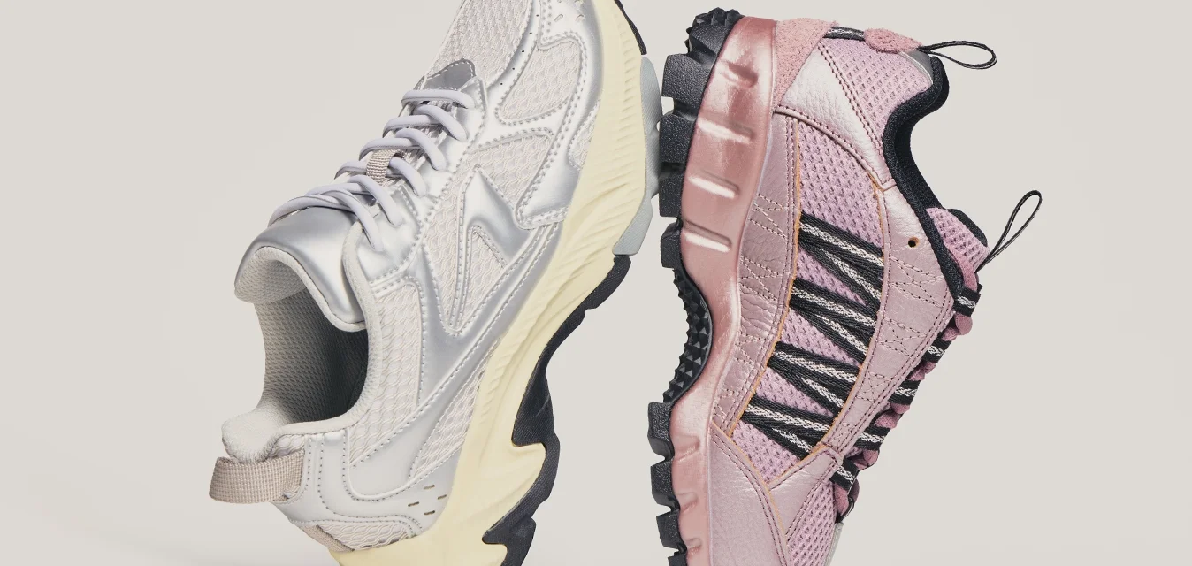 Een paar sportieve schoenen, de linker in metallic zilver met een crèmekleurige zool, de rechter in roze met zwarte veters, staan zool-op-zool op een neutrale achtergrond.