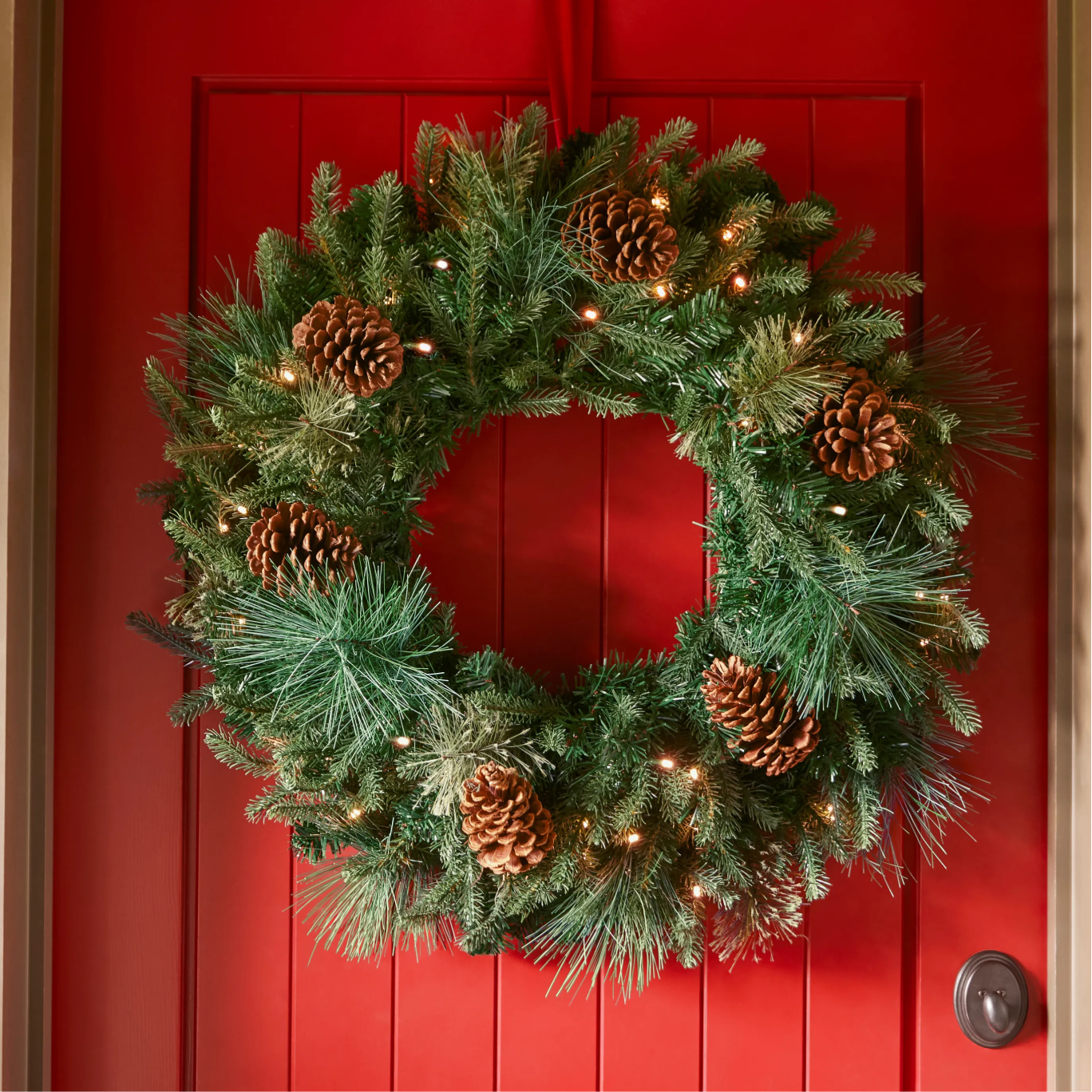 Pre-lit Christmas wreath with pinecones and mixed greenery hanging on a red front door.