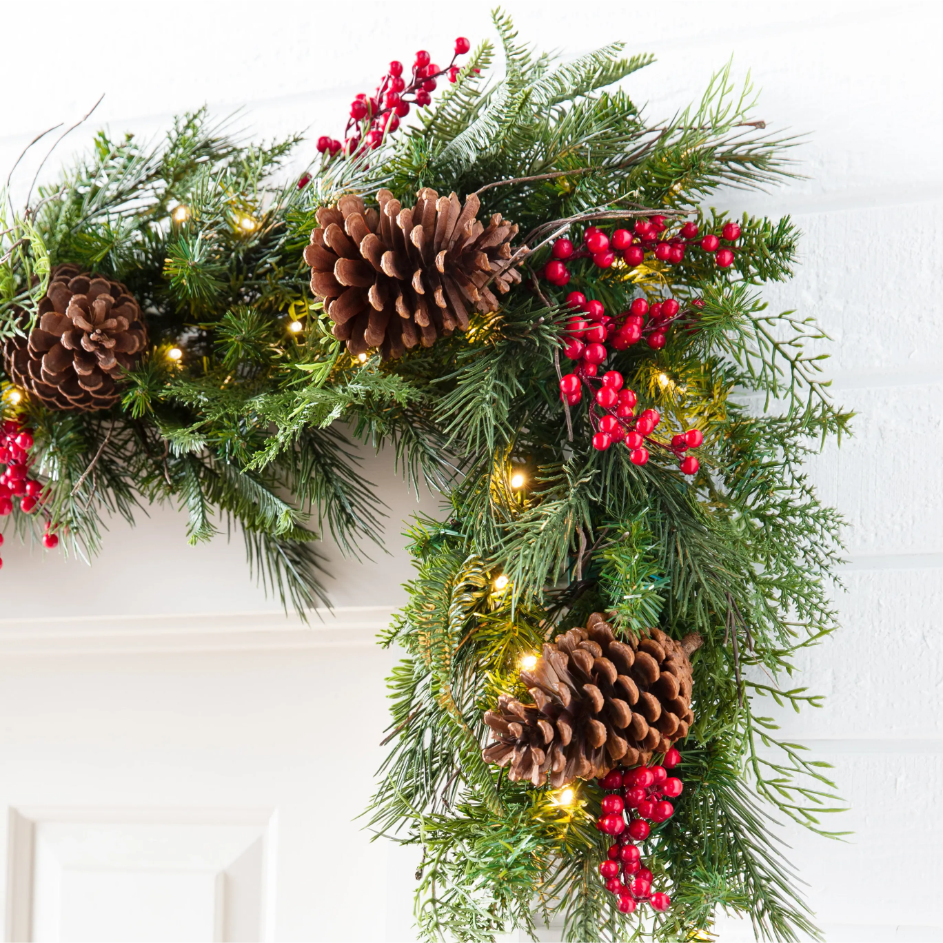Pre-lit Christmas garland with pinecones, red berries, and mixed greenery draped over a white mantel.