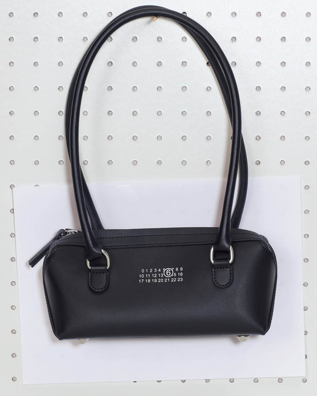 Black leather shoulder bag with long double straps and silver hardware, barrel-shaped with small white numeric logo, on pegboard background