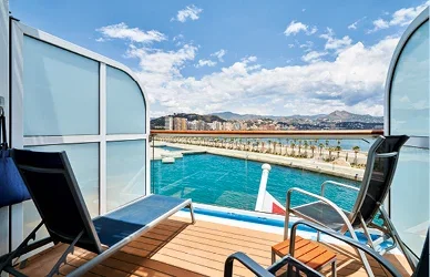 Private balcony with loungers overlooking a cruise ship pool, marina and distant mountains.