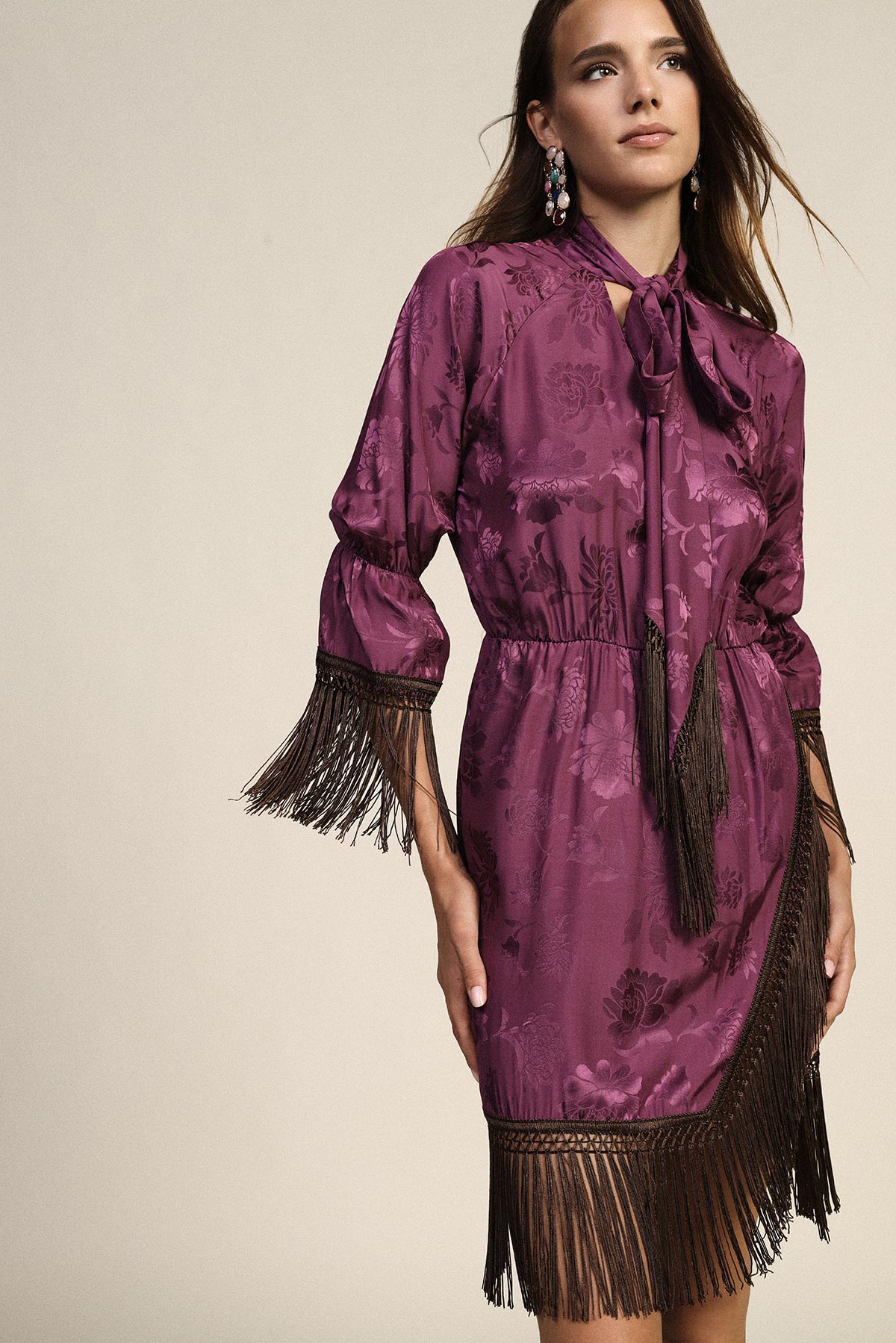 Woman wearing purple floral silk dress with bow neckline and black fringe trim, statement earrings on neutral background