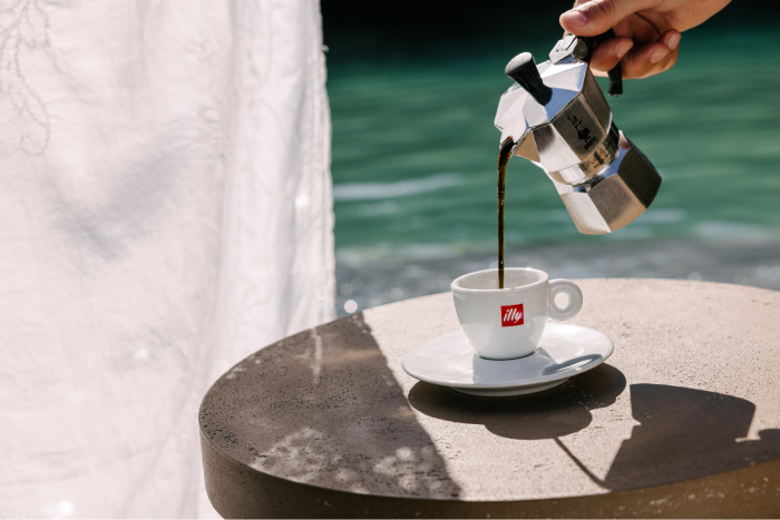 Fresh coffee being poured from a moka pot into an illy espresso cup on a sunlit outdoor table