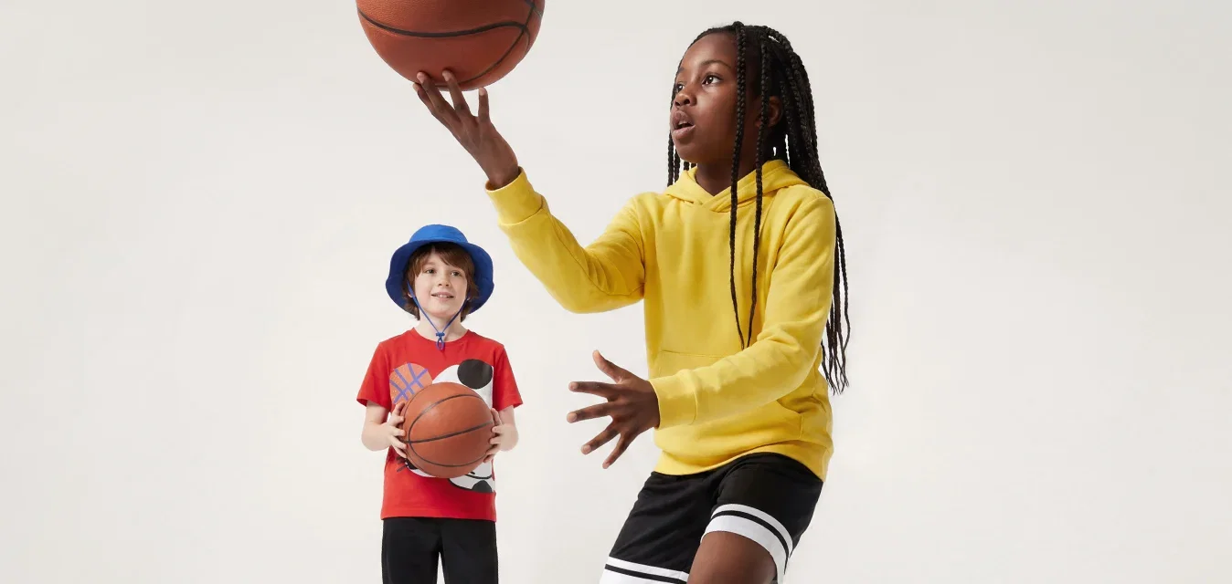Zwei Kinder spielen mit Basketbällen, eines in einem gelben Hoodie dribbelt, das andere in einem roten Hemd und blauer Mütze steht still.