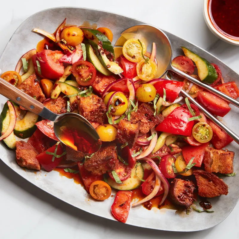 Chili crisp vinaigrette being poured over a platter of tomato and zucchini panzanella.