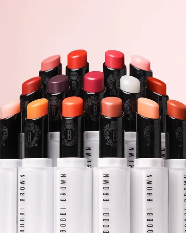 A semi-circular arrangement of colourful Extra Lip Tinted Balms in various shades, against a pastel pink background.