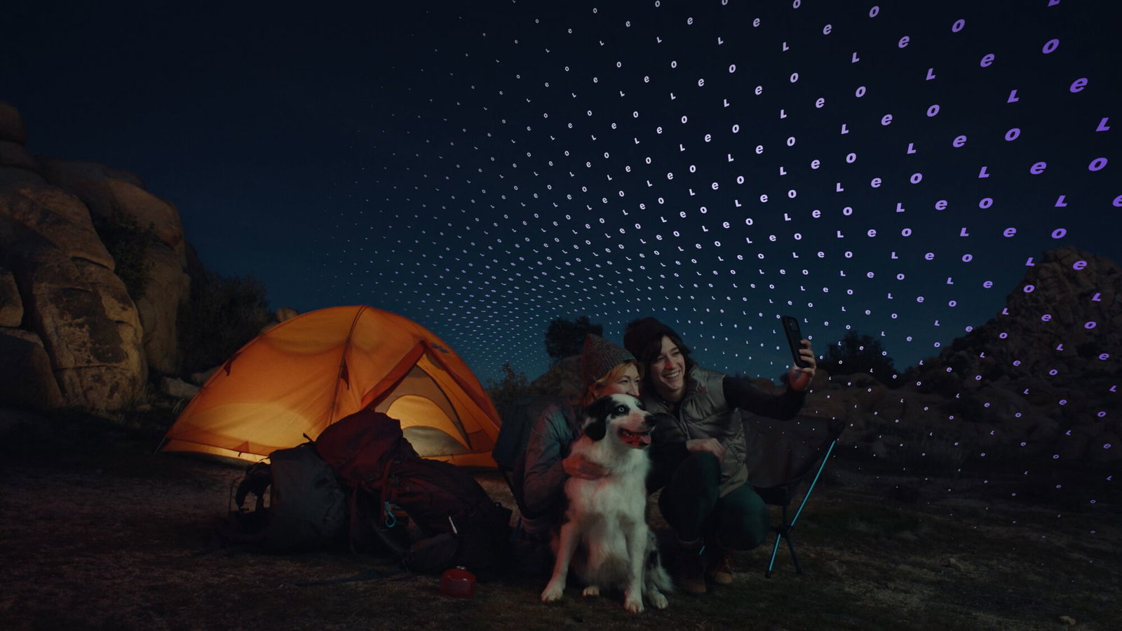 Friends and their dog pose by an illuminated orange tent as a digital canopy of letters sweeps across the night sky.