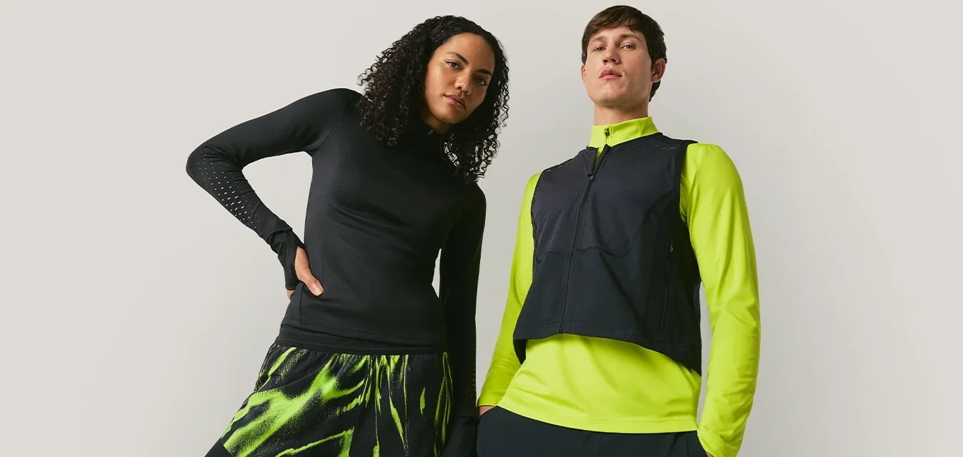 Dos modelos posan frente a un fondo liso, uno viste ropa deportiva negra con verde neón, y el otro lleva una camiseta negra y una chaqueta verde neón con chaleco.