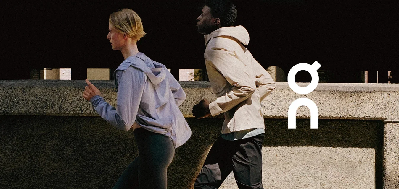 Dos personas corriendo al aire libre, vistiendo chaquetas deportivas, leggings y zapatillas de correr en una calle urbana soleada.