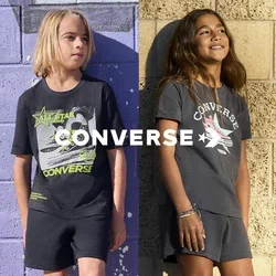 Dos niños con camisetas gráficas de Converse y pantalones cortos están de pie contra paredes texturizadas, una morada y una beige, sonriendo y mirando hacia otro lado.
