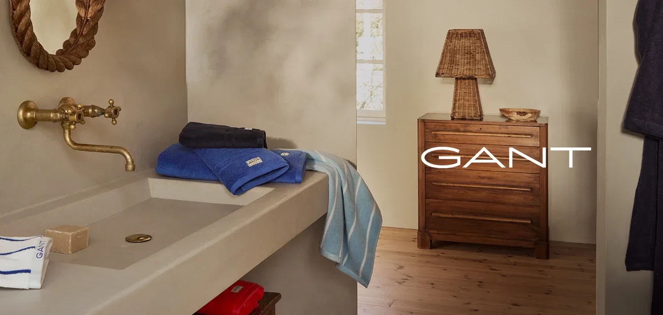 Badkamer met beige muren, een gebogen touwen spiegel, een messing kraan, opgestapelde handdoeken (blauw, marineblauw en gestreept), een houten dressoir en een geweven lamp.