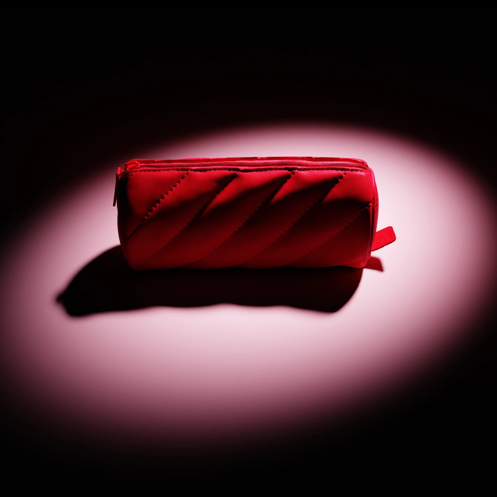 A vibrant red quilted cylindrical makeup pouch highlighted under a central spotlight on a dark background.