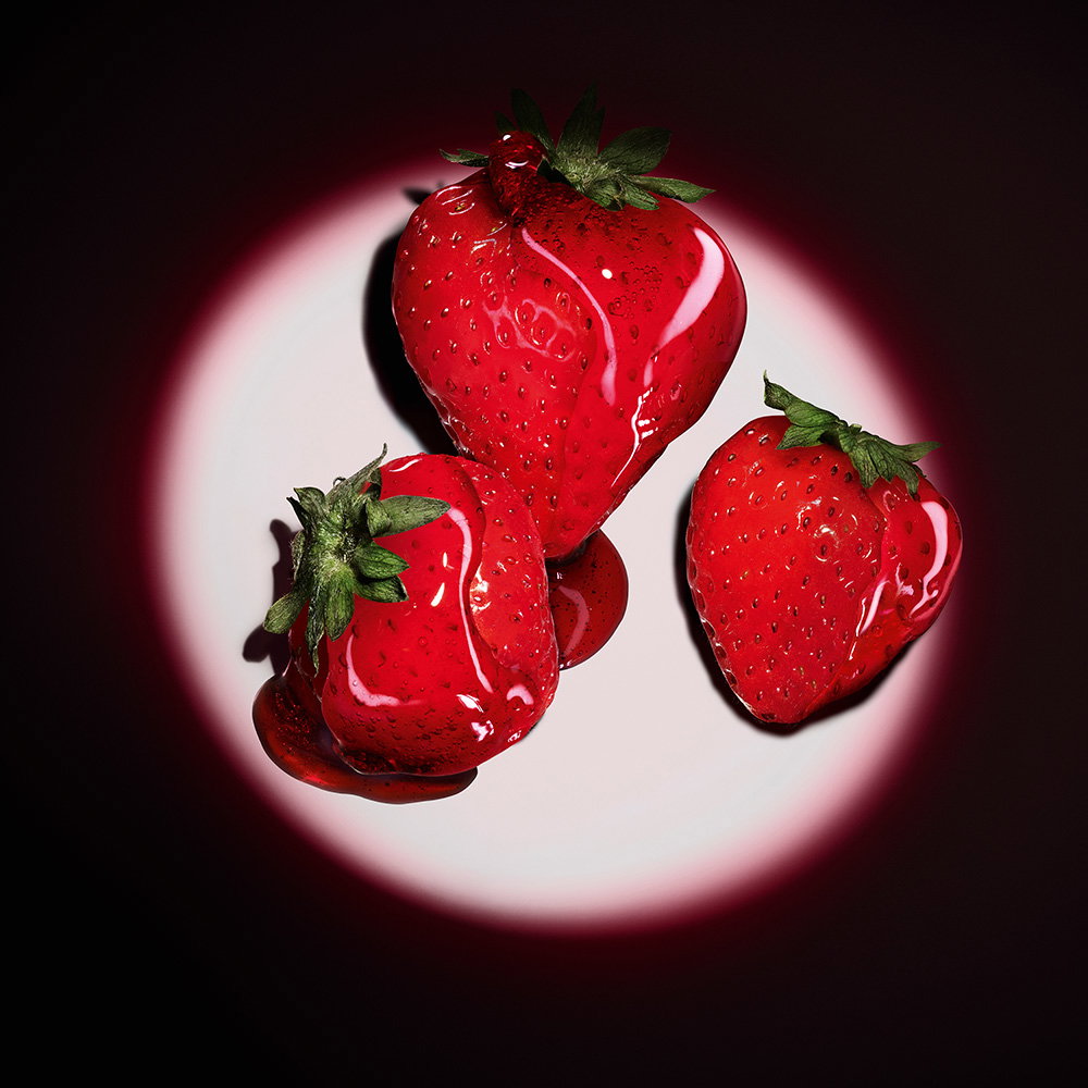 Three fresh red strawberries glazed with a glossy red syrup under a circular spotlight.