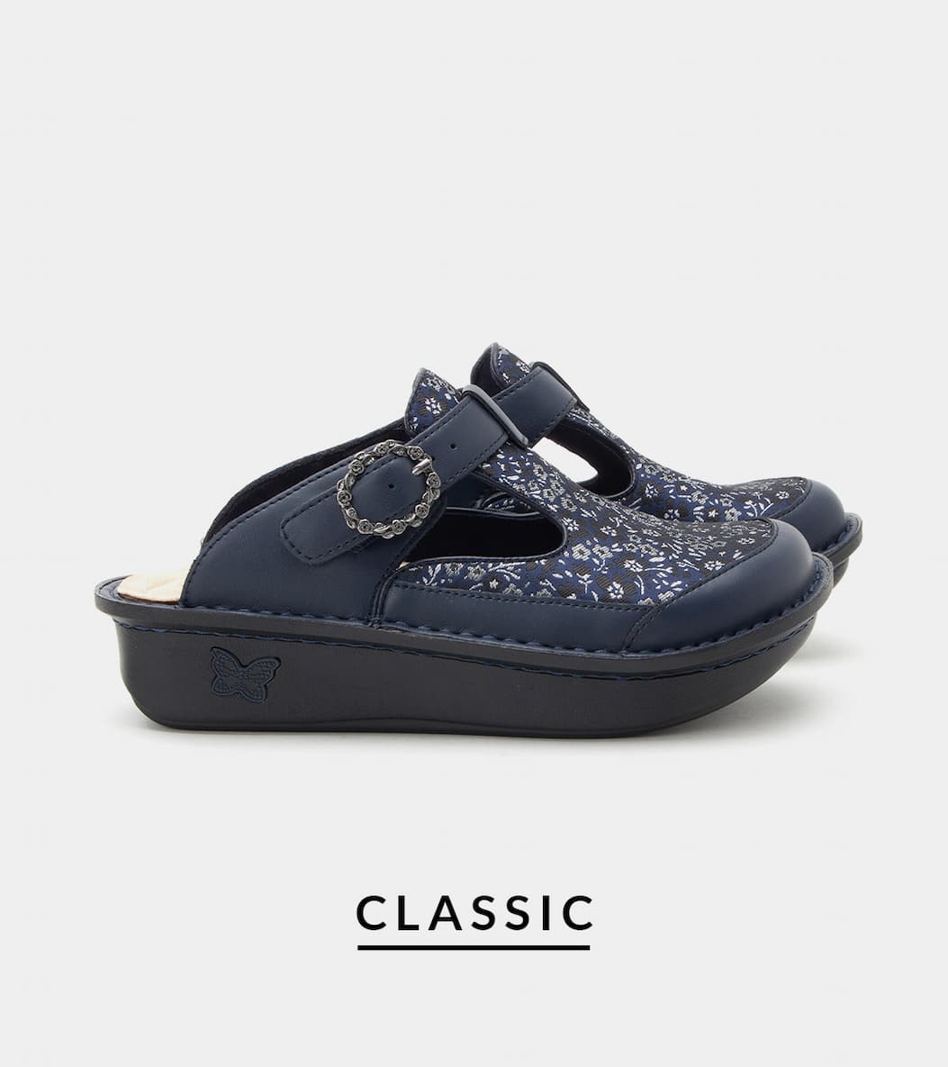Pair of dark navy blue clog-style slip-on shoes with a silver floral print upper, an instep strap with an ornate round silver buckle, and a thick black sole featuring a debossed butterfly detail. Product name "CLASSIC" printed beneath on a light gray background.