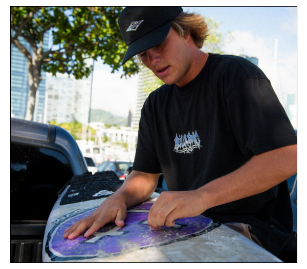 Billabong surfer waxing his surfboard wearing a hat and a t-shirt.