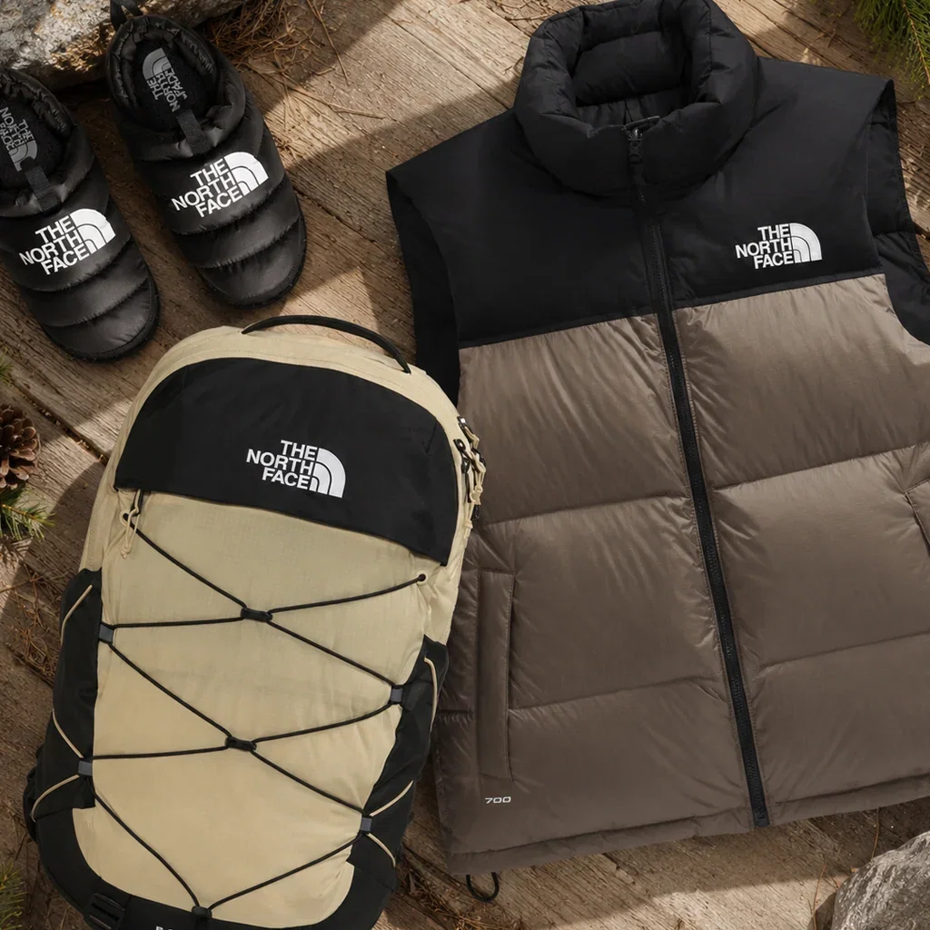 The North Face gear collection featuring cream and black backpack, black and gray puffer vest, and black winter boots on wooden surface.