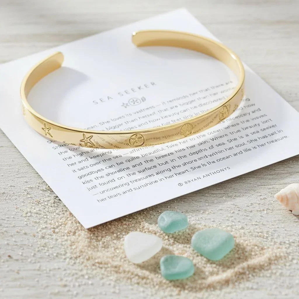 Gold cuff bracelet with sea-themed engravings displayed on inspirational card with sea glass stones