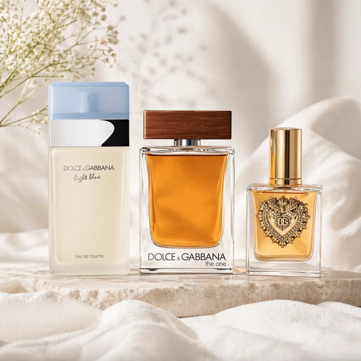 Three Dolce & Gabbana fragrances displayed on stone: Light Blue, The One, and Devotion with dried flowers.