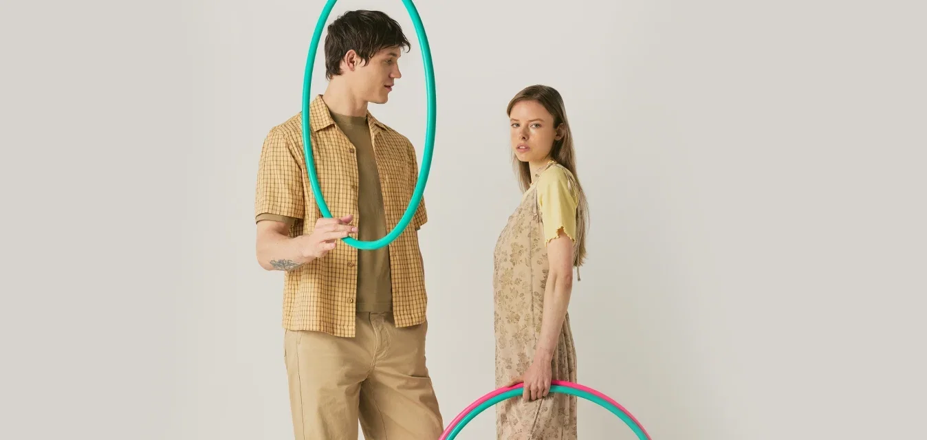 Un hombre y una mujer están de pie sosteniendo aros hula hoop coloridos; él sostiene un aro turquesa cerca de su rostro, ella sostiene dos aros a su lado.