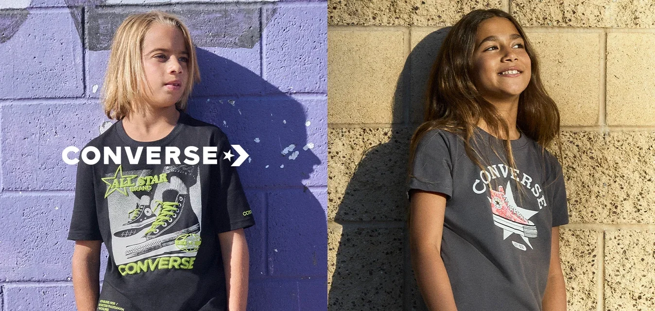 Dos niños con camisetas gráficas de Converse y pantalones cortos están de pie contra paredes texturizadas, una morada y una beige, sonriendo y mirando hacia otro lado.