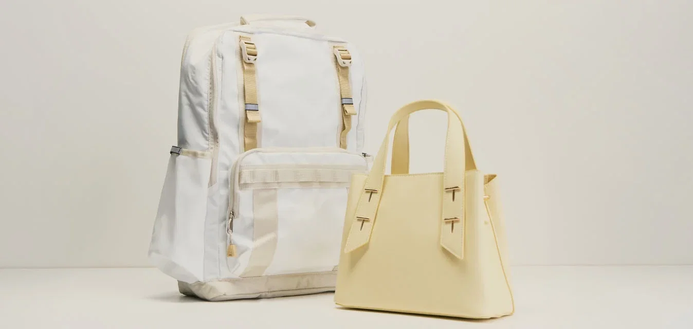 Mochila blanca con correas beige junto a un pequeño bolso beige sobre un fondo claro y liso, ambos de pie.