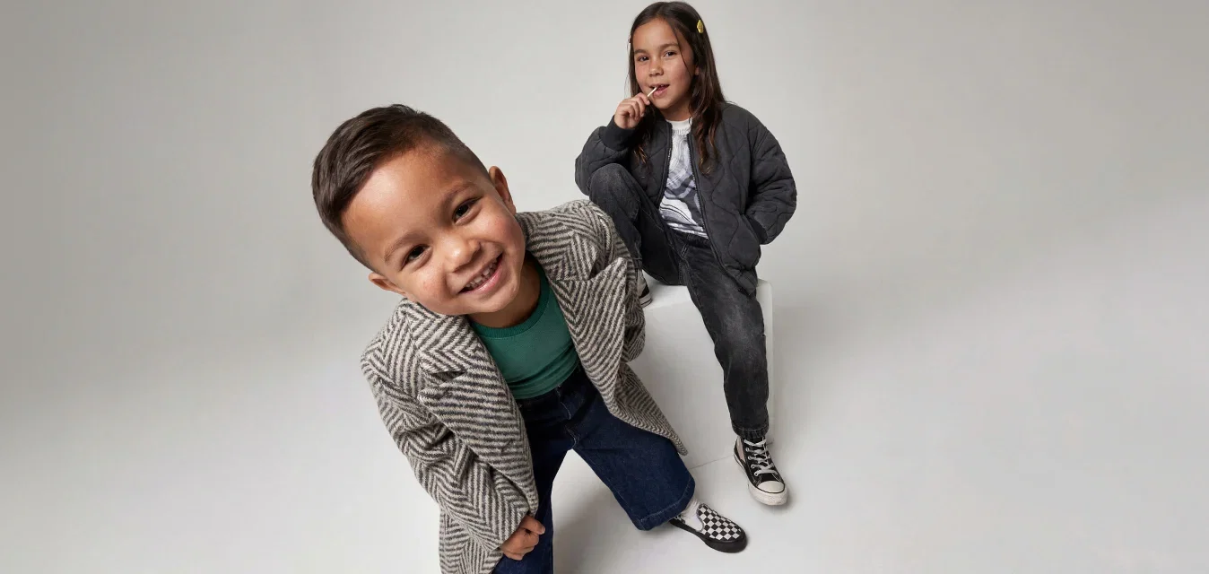 Un niño sonriendo con un abrigo de espiga se inclina hacia adelante mientras una niña con una chaqueta negra se sienta detrás de él, ambos sobre un fondo liso y claro.