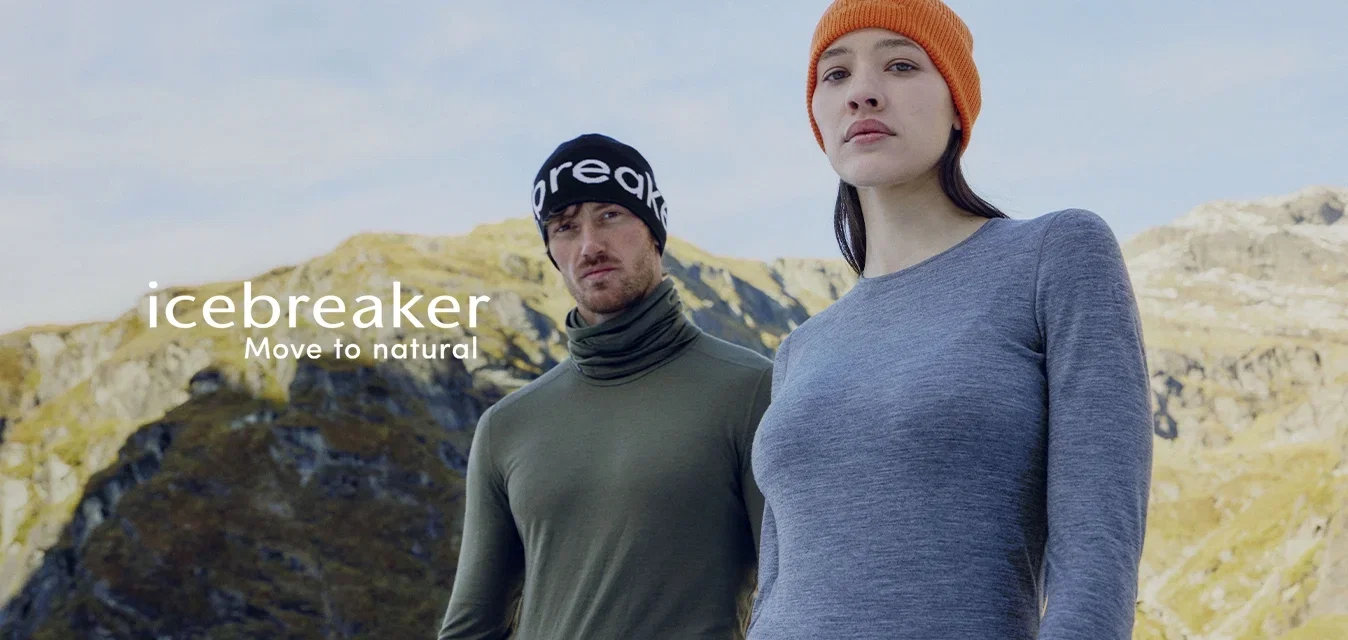 Twee volwassenen in buitensportkleding met merkgebonden baselayers en mutsen staan voor een bergachtig landschap onder een heldere hemel.