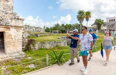Visitors walk through the ancient Mayan ruins of Tulum, surrounded by stone structures, tropical plants and palm trees under a bright blue sky.