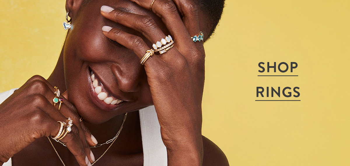 Shop rings. Smiling woman wearing multiple gold rings against a yellow background.