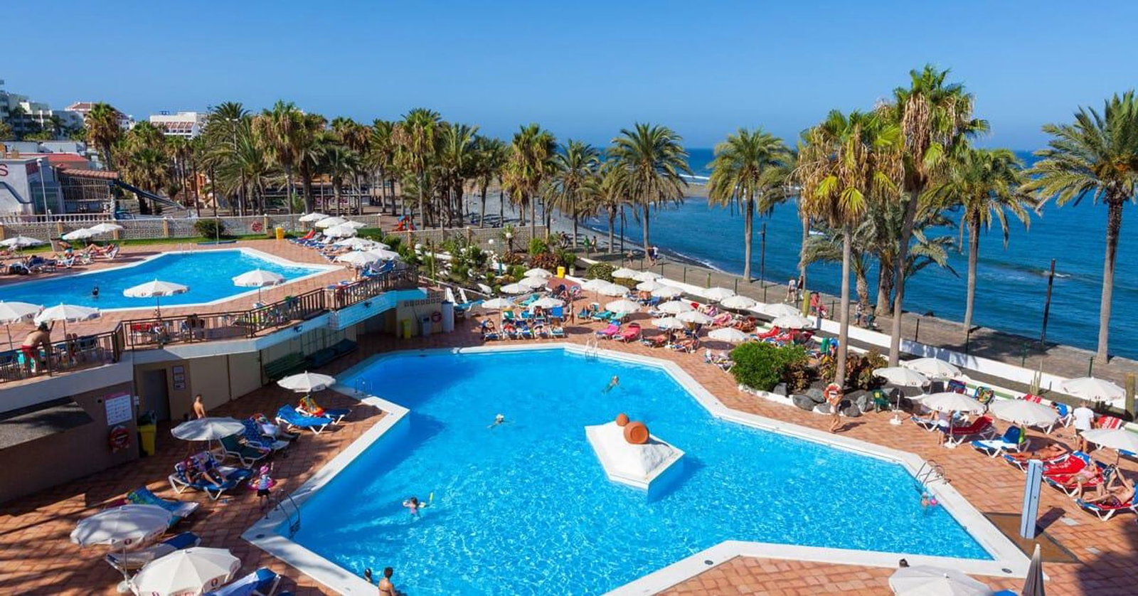 Resort swimming pools and sun-loungers beside palm trees on a seaside promenade, clear blue water and umbrellas