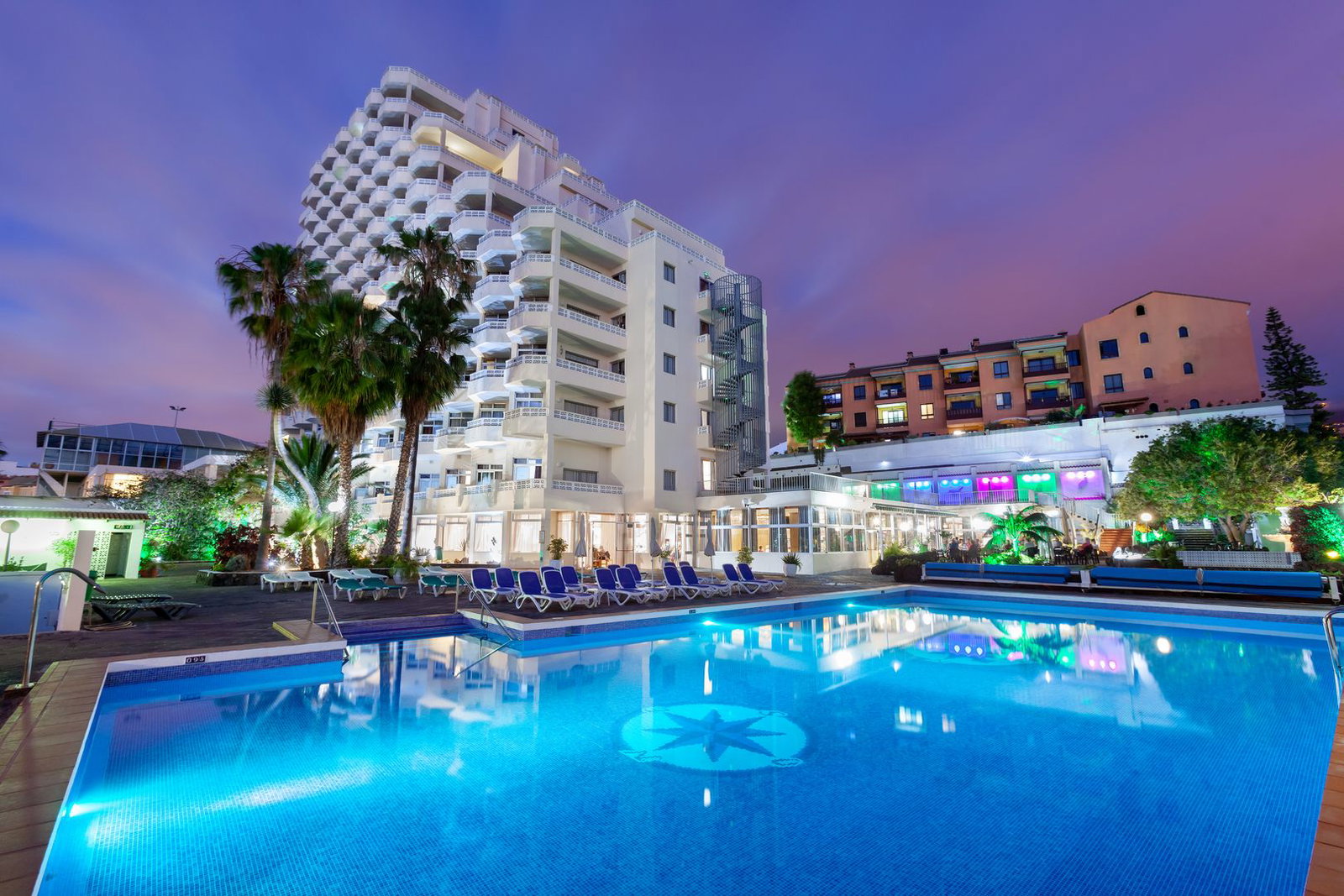 Illuminated hotel with balconies and palm trees reflected in a lit outdoor swimming pool with loungers