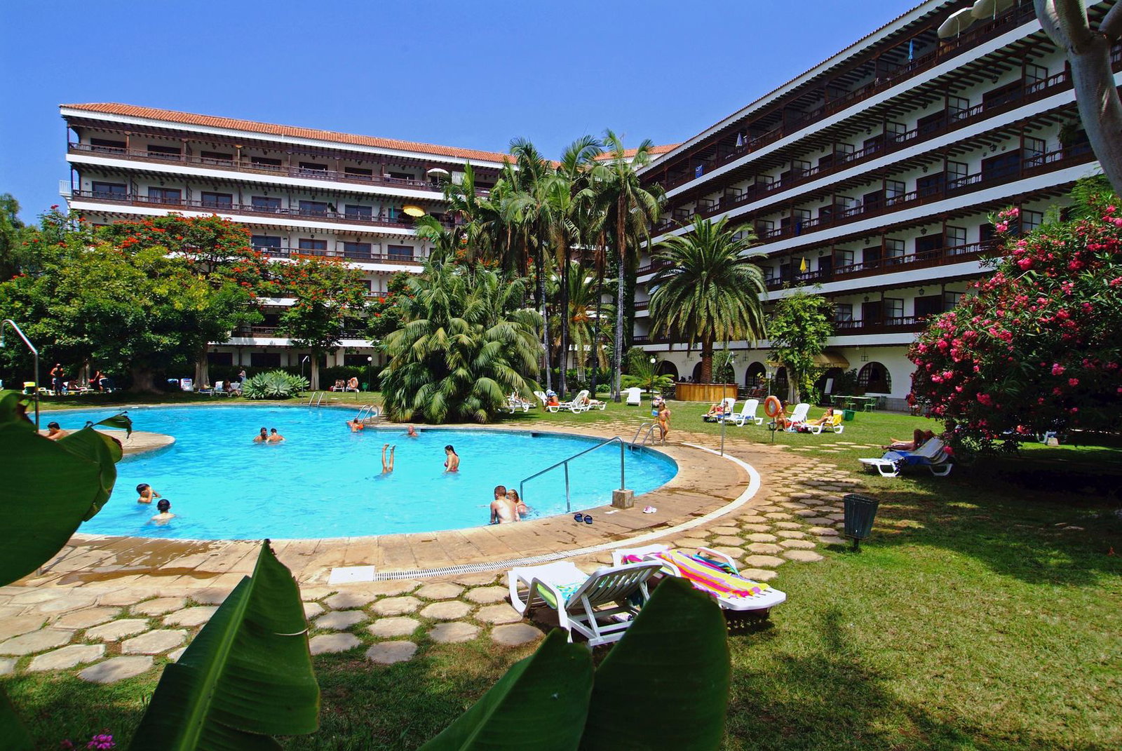 Sunny resort hotel courtyard with circular outdoor swimming pool, palm trees and sunbeds, guests swimming and relaxing