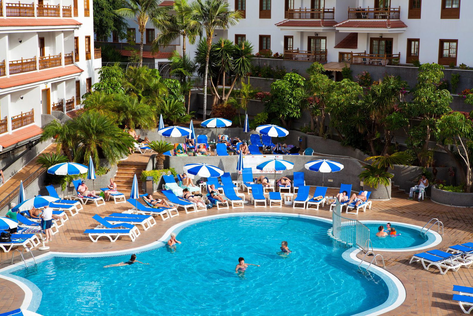 Hotel resort swimming pool and poolside with blue sunbeds and umbrellas, palm trees and guests relaxing