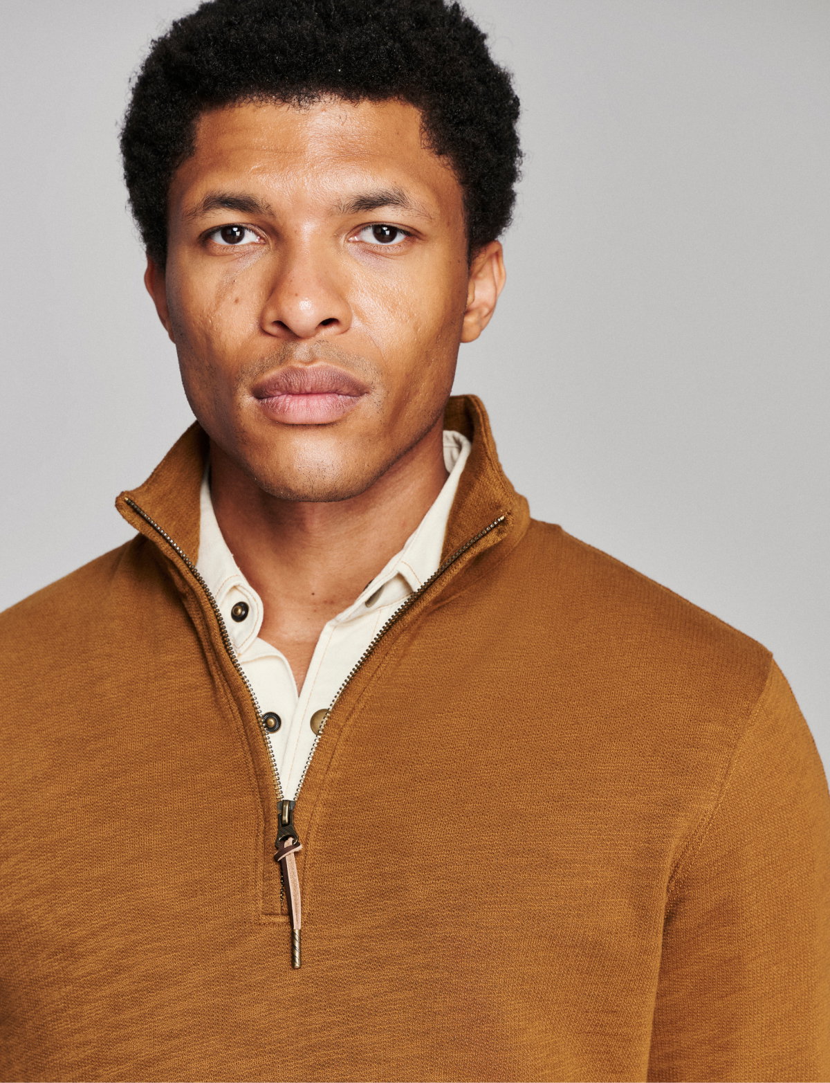 Model wears a brown half zip over a neutral button up on a grey background.