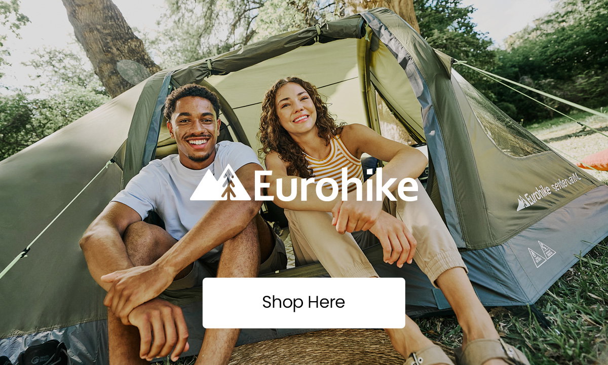 Two adults sitting at the entrance of a green camping tent in a wooded outdoor setting, with “Eurohike” logo overlay and a “Shop Here” button centered on the image.