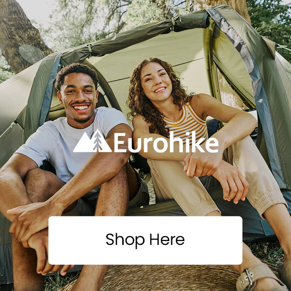 Two adults sitting at the entrance of a green camping tent in a wooded outdoor setting, with “Eurohike” logo overlay and a “Shop Here” button centered on the image.
