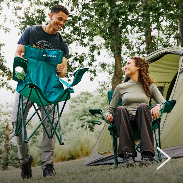 Two adults setting up folding camping chairs on grass next to a pitched tent under trees, with one person holding a folded chair while the other is already seated in a matching chair.