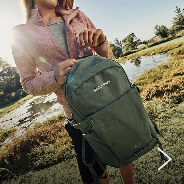 Close-up of a person adjusting the zip of a green Eurohike backpack beside a small pond, with grassy ground and bright sunlight in the outdoor setting.