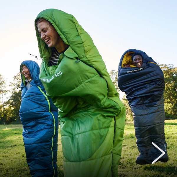 Three adults standing outdoors wrapped inside full-body sleeping bags in blue, green, and black, clustered together on grass in daylight.