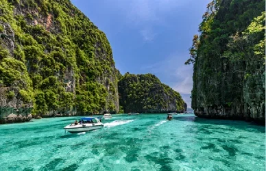 Towering limestone cliffs rise above crystal-clear waters, with boats cruising through a tropical lagoon in Thailand.