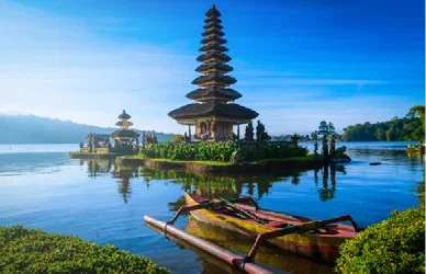 Traditional Balinese temple buildings sit on a peaceful lake, with a wooden boat in the foreground and lush greenery in the background, Indonesia.