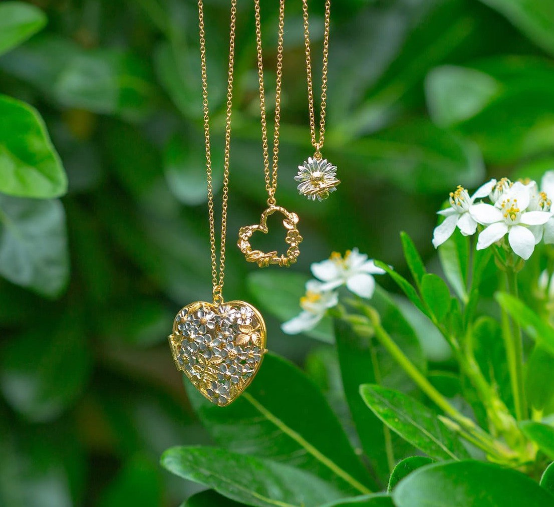 Gold bee, daisy and floral-inspired necklaces hanging amongst white summer flowers and green foliage.