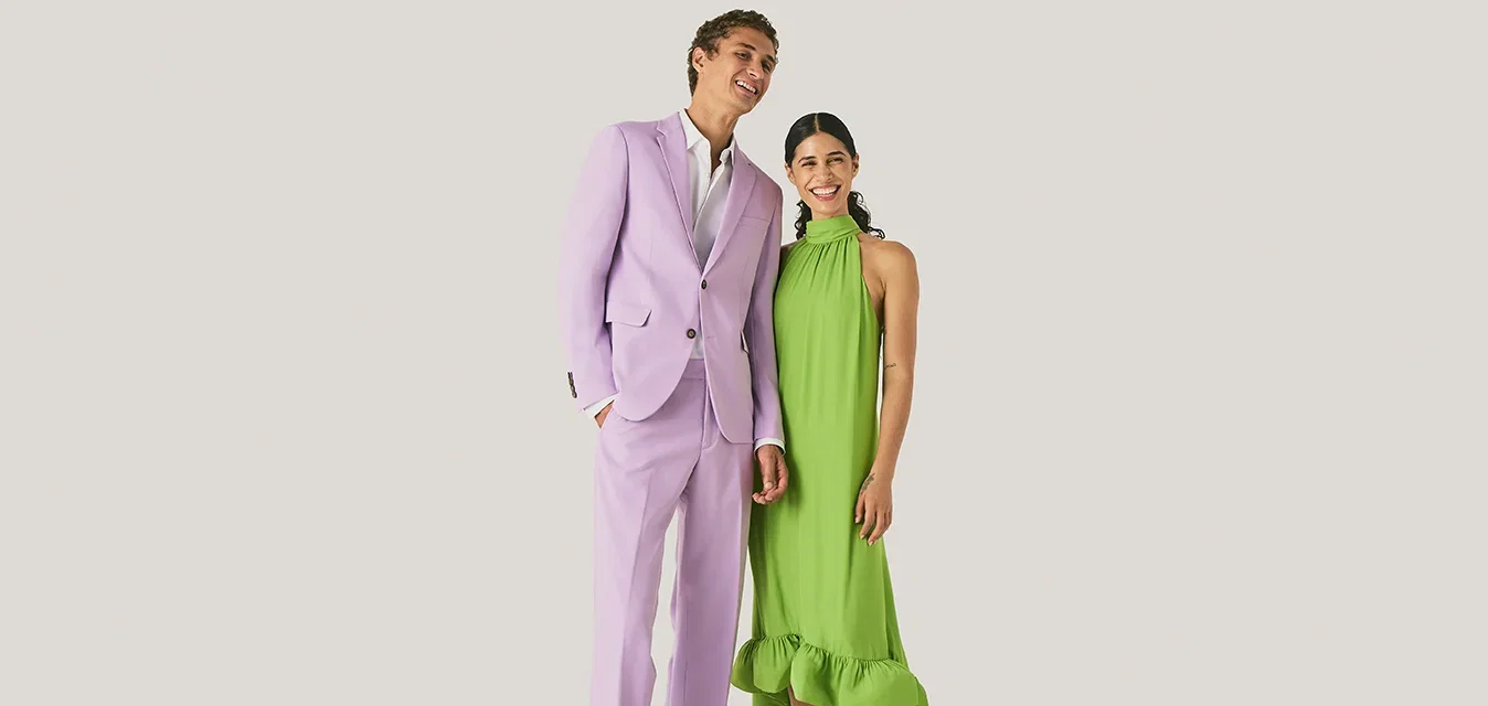 Un homme en costume violet clair et une femme en robe sans manches vert vif sourient debout devant un fond clair uni.